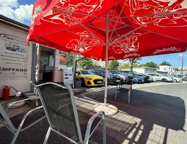Parkovisko Parkparadies – Na parkovisku stoja zaparkované autá. Vedľa sú stoličky a stolík pod červeným slnečníkom. Areál je otvorený na slnko.