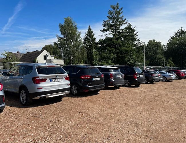 Parking Park In Fly Out - Samochody stoją na szutrowym placu. W tle widoczne są drzewa i ogrodzenie. Pogoda jest słoneczna.