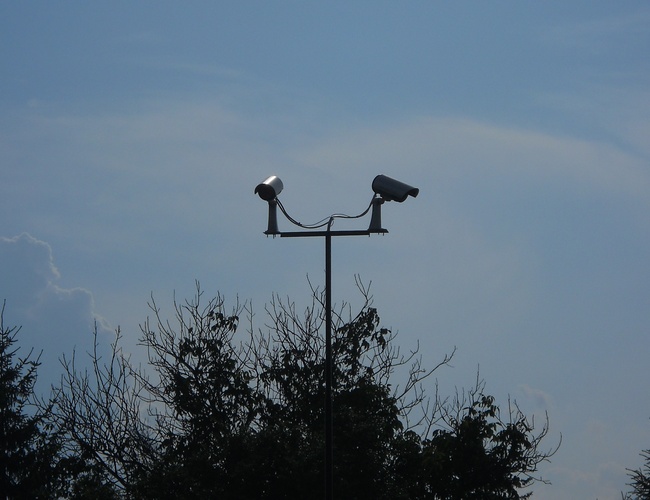 Parkovisko Auto Park – na obrázku sú dve kamery na stĺpe. Kamery monitorujú okolie parkoviska. V pozadí je vidieť korunky stromov proti oblohe.