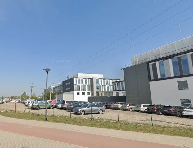 Parkovisko Piątka - Otvorený priestor so zaparkovanými autami a oplotením. Vedľa sa nachádzajú moderné budovy.