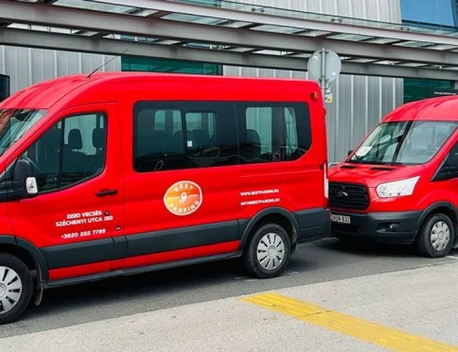 Dos furgonetas rojas de transporte con el logo del aparcamiento visible en las puertas. Aparcadas junto a la terminal de salidas del aeropuerto.