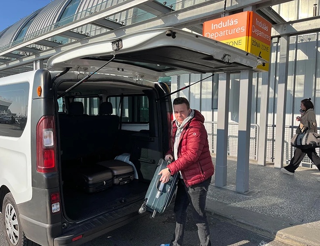 Osoba wkładająca bagaże do otwartego bagażnika busa przy strefie odlotów pod zadaszeniem terminala.