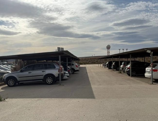 Szeroki parking z kilkoma zaparkowanymi autami pod zadaszonymi miejscami, w tle więcej zaparkowanych samochodów pod zadaszeniem i na utwardzonym terenie.