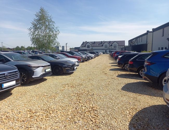 AutoParking24h – Szutrowy parking z zaparkowanymi samochodami po obu stronach. W tle są budynki i drzewo.