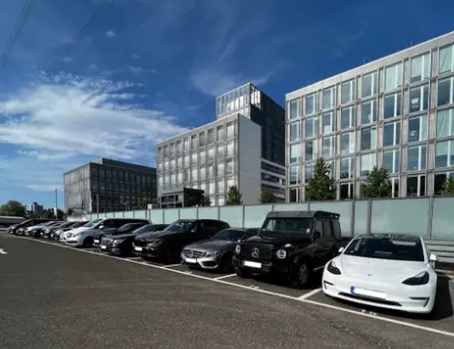 Parkovisko Park2Travel Valet Outdoor - Autá stoja v rade na otvorenom parkovisku. Pozadie tvoria moderné kancelárie a modrá obloha.