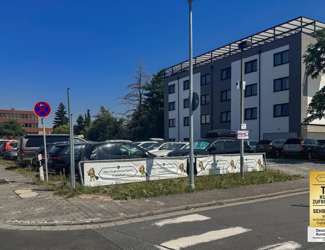 Parking Mein-Flughafenparkplatz-Frankfurt P3. Widok z ulicy na parking przed budynkiem mieszkalnym, w tle zaparkowane samochody i znak drogowy.