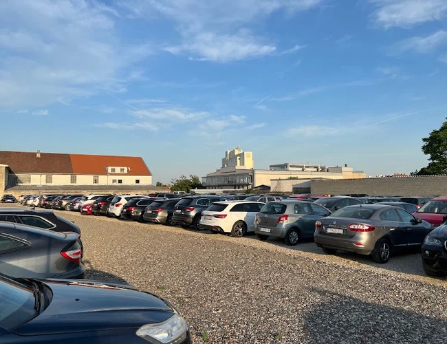 Parqueo de Aeropuerto Parktiger 2 - Hay coches estacionados en el aparcamiento. El terreno es de tierra y espacioso. Al fondo hay edificios.