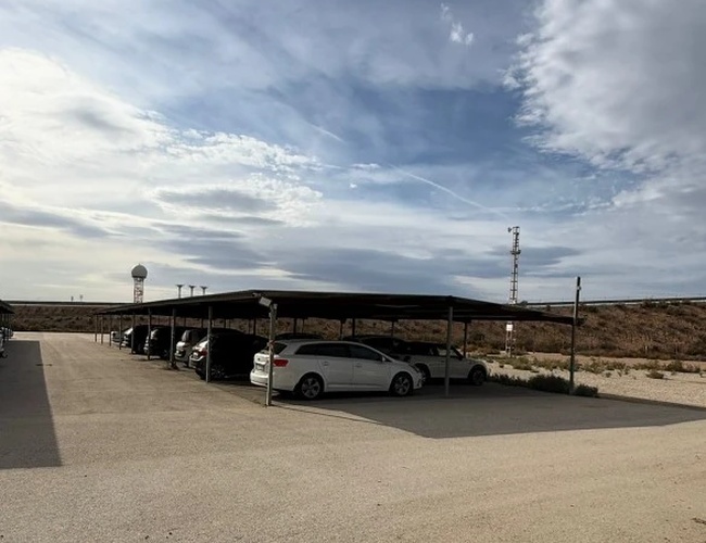 Parking z kilkoma zaparkowanymi autami pod zadaszonymi miejscami, w tle więcej zaparkowanych samochodów pod zadaszeniem i na utwardzonym terenie.