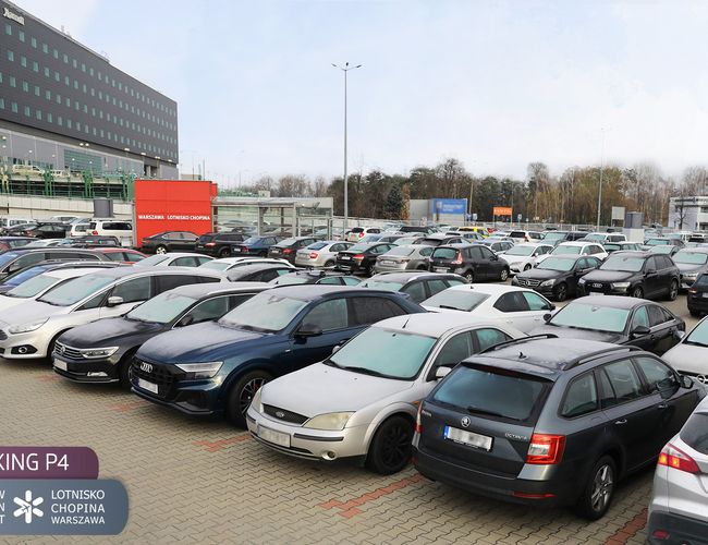Oficjalny Parking lotniska P4 – widoczne są zaparkowane samochody. Obok stoi duży czarny budynek. Teren jest wyłożony kostką brukową.