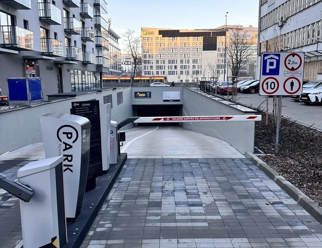 Najvyššia parkovacia garáž – podzemný vstup s rampou. Vedľa stoja budovy a dopravné značky. Parkovisko vyzerá byť moderné a priestranné.