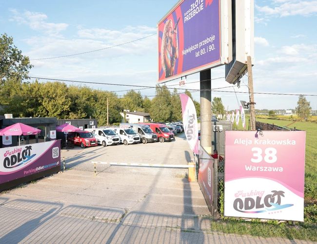 Parkoló Elindulás - A kavicsos felületen sok autó áll. A parkoló kerítéses. Távolabb az út és az utcai lámpák láthatók.