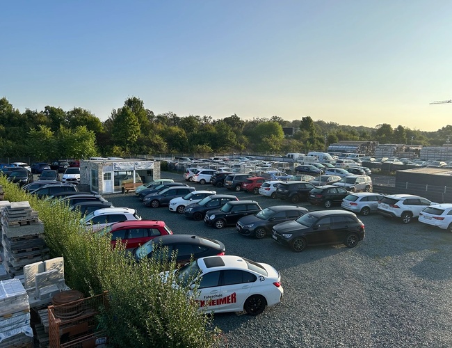 Estacionamiento de Valet de Transporte – El estacionamiento es de grava y está cercado. Hay muchos coches en el recinto. En el fondo se pueden ver árboles.