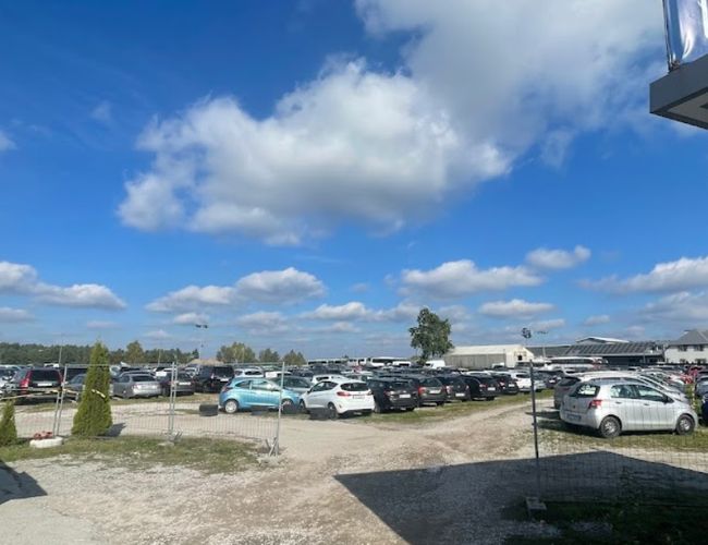 Aparcamiento bajo el molino - Aparcamiento lleno de coches, cercado con malla. En el cielo se ven nubes. Al lado hay un edificio gris con un letrero.