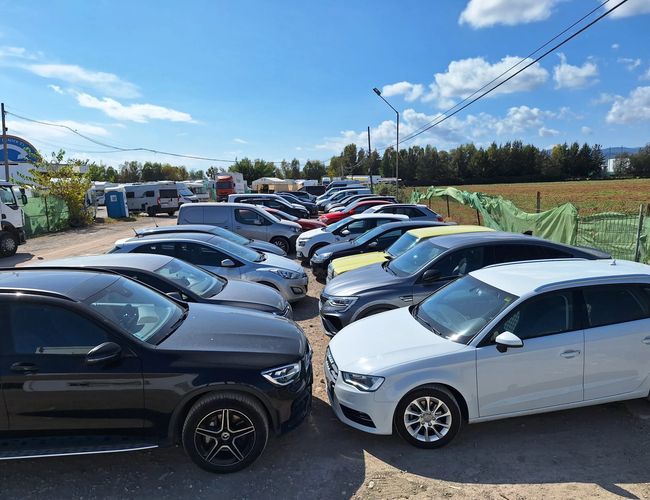 Parking Low Valet - Kilkanaście aut stoi na żwirowym placu. Obok widoczne są krzewy i ogrodzenie. Na horyzoncie rosną drzewa.