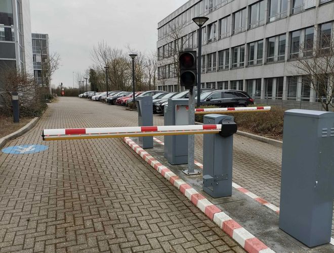 Sun Park Airport Parking Outdoor – Wjazd z szlabanem i sygnalizacją świetlną. Obok stoi rząd zaparkowanych samochodów. Teren jest utwardzony.
