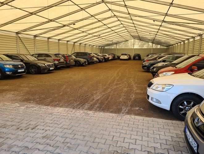 B1 Parking zadaszony - Pod dachem stoją zaparkowane samochody. Teren ma nawierzchnię z kostki brukowej.