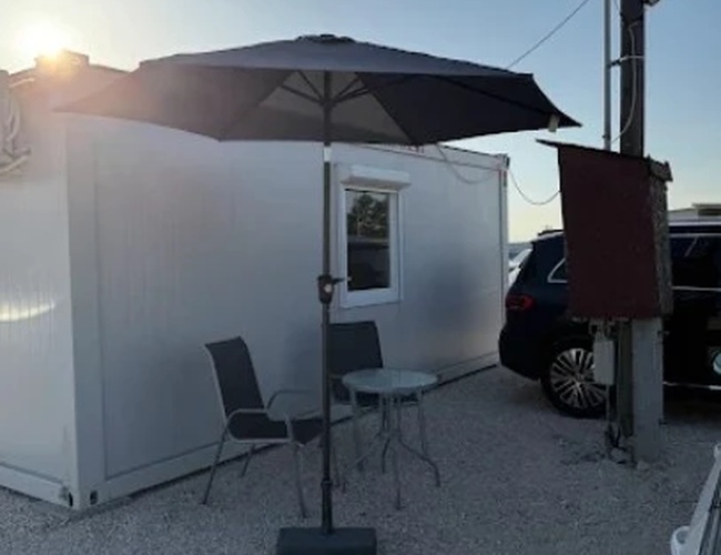 Niewielki biały kontener biurowy z parasolem i dwoma krzesłami przed wejściem, oświetlony niskim słońcem.