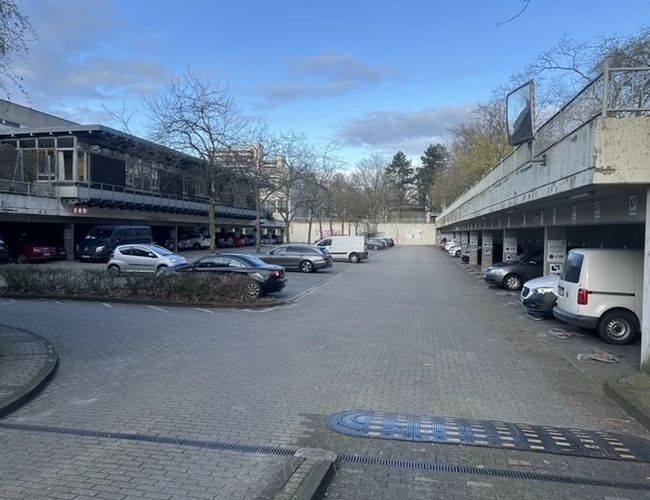 Valet Parking DUS - Parking zadaszony, z zaparkowanymi samochodami po obu stronach. Obszar jest otoczony drzewami, niebo jest niebieskie.