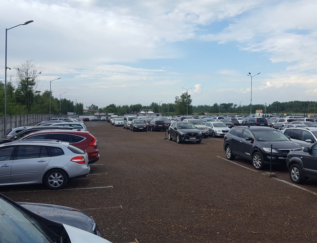 AAP-66 Parking – duży, otwarty teren z licznymi samochodami. Parking znajduje się obok ogrodzenia z zielenią w tle. Wetter wygląda na pochmurny, a latarnie są zamontowane.