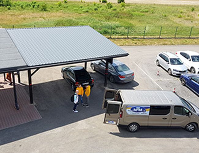 Parking MTM - Na parkingu stoją zaparkowane samochody. Dwa samochody są pod zadaszeniem. Na parkingu widoczne są osoby.