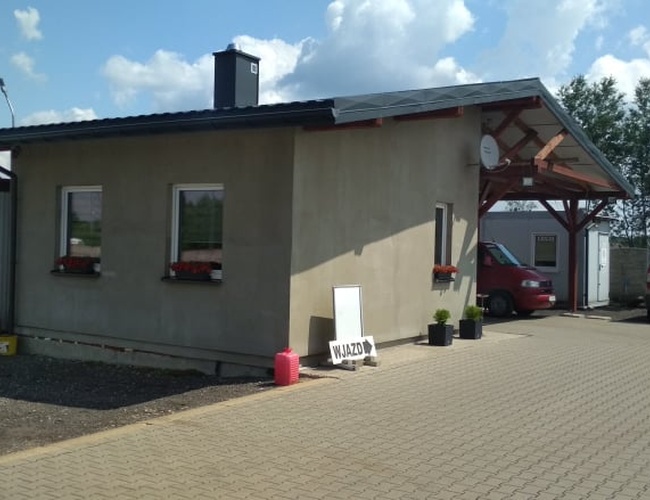 ESTACIONAMIENTO de TRAVEL - Edificio con toldo en la entrada. Al lado hay un coche. Todo está rodeado de una cerca con vegetación.