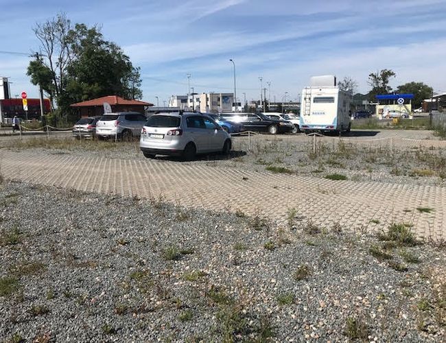 Auto Airport Samoobsługowy – Parking z kostką brukową i żwirem. Kilka aut zaparkowanych na wyznaczonych miejscach. Teren otoczony niskim ogrodzeniem.