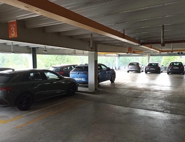 Półotwarty parking z miejscami postojowymi pod zadaszeniem. Samochody zaparkowane są wzdłuż drogi przejazdowej, a na końcu parkingu widoczne jest światło dzienne.
