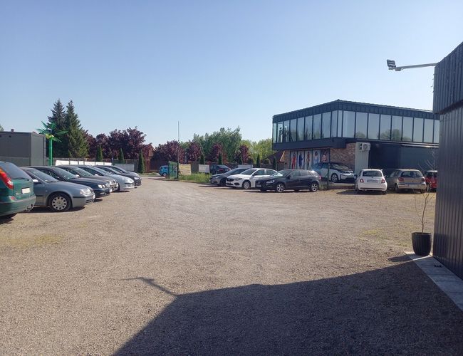 AutoParking24h - Na żwirowym placu stoi wiele aut. Po prawej stronie jest przeszklony budynek. Teren jest otoczony roślinnością.