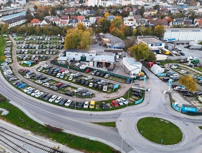 Parktiger Flughafenparkplatz 1 – Große Parkfläche mit vielen Autos. Das Gelände ist eingezäunt und gut organisiert. In der Nähe befinden sich ein Kreisverkehr und Gebäude.