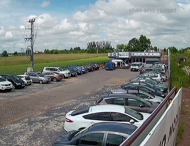 PARKING P24 PREMIUM - Na parkingu stoją samochody. Teren jest ogrodzony i utwardzony. W tle widać niewielki budynek.