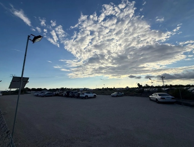 Duży parking o zmierzchu z rzędem zaparkowanych aut, latarnia uliczna i efektowne chmury na niebie.