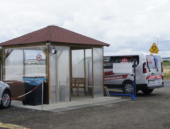 Aparcamiento Aeropuerto 24h - Pequeño pabellón de madera con techado. Al lado hay una furgoneta blanca. El terreno está pavimentado y cercado.