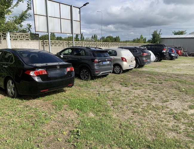 A1 Parking Max - Niekoľko áut stojí na námestí. Pozemok je ohradený betónovým plotom. Vedľa vidieť lampu a stromy.