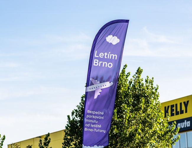 Parking Letim Brno – na zdjęciu widoczna jest fioletowa flaga reklamowa. Znajdują się na niej napisy informacyjne oraz grafika samolotu. W tle widać drzewo i fragment budynku.