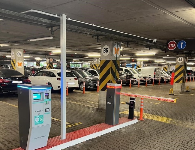 Parkovisko P2 - Okęcie Sever Multifunkčné - viditeľné sú zaparkované automobily a závoru. Uprostred sa nachádza platobný automat. Steny a stĺpy sú označené.