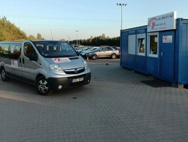 Pierwszy Parking - Widoczny jest niebieski kontener i samochód. W tle stoją zaparkowane auta.
