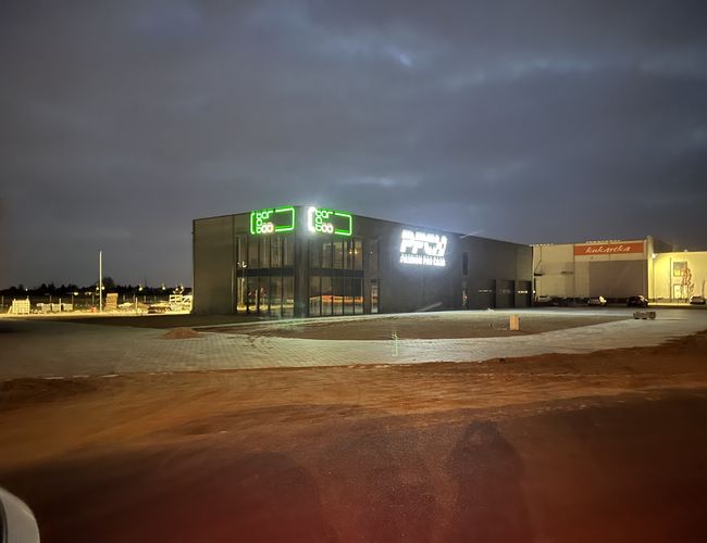 Q-Parking – Budynek z neonowym napisem świeci w nocy. Plac jest brukowany i nieoświetlony. Tło stanowią magazyny.