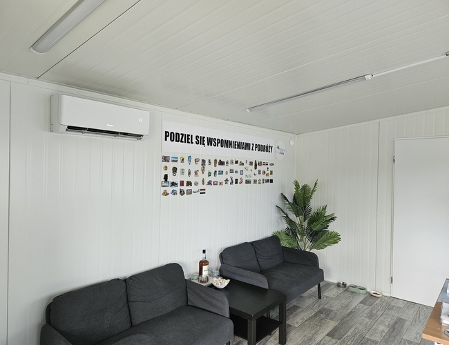 Holiday Parking - Wnętrze poczekalni z sofą i stolikiem. Na ścianie wiszą magnesy z podróży. Roślina stoi w rogu pokoju.