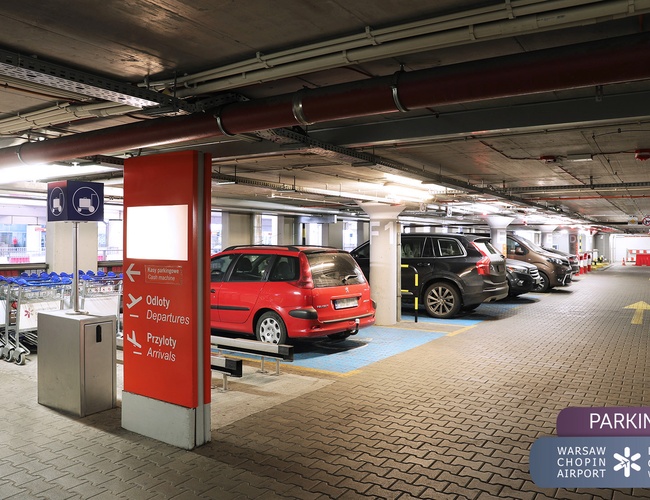 Oficiálne parkovisko Letiska P2 - Podzemné parkovisko s autami. Viditeľné smerové značky a košíky na batožinu. Stropné osvetlenie.