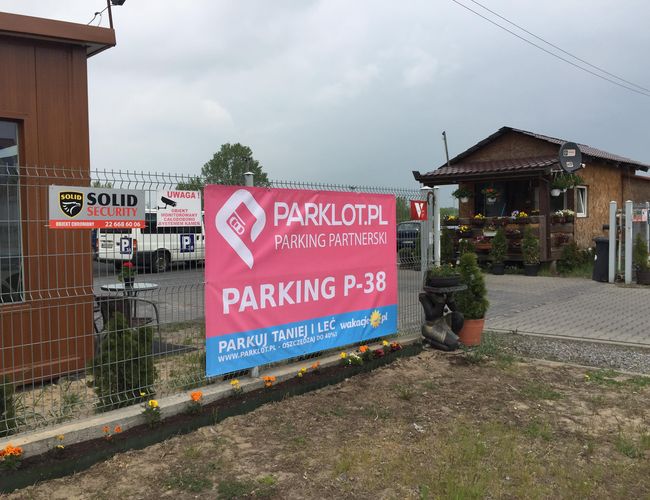 Estacionamiento P38 - El estacionamiento está cercado con una malla. Al lado hay un edificio de madera. Frente a él crecen flores en macetas.
