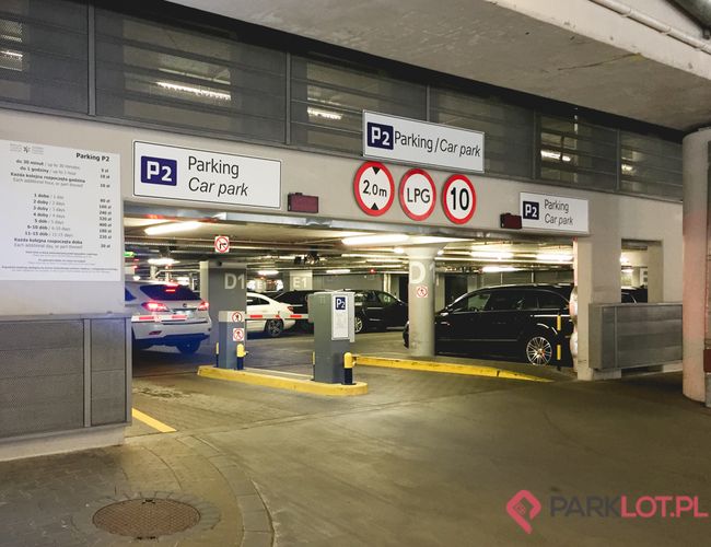 Oficjalny Parking Lotniska P2 – Kryty parking z wjazdem dla aut. Znaki wskazują ograniczenia wysokości, LPG i prędkości. W środku zaparkowane samochody.