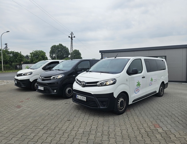 Holiday Parking – trzy pojazdy stoją obok siebie. Dwa białe mają logo z palmą. Parking jest brukowany i ogrodzony.