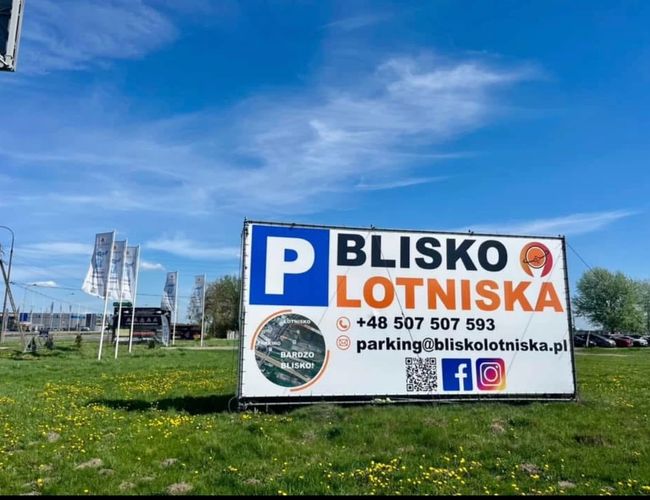 Parking Blisko Lotniska – Rząd samochodów stoi na żwirowym placu. Teren jest otwarty, z widocznymi flagami reklamowymi. W tle widać drzewa.