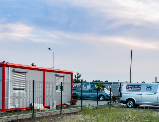 AAP-66 – Parking jest ogrodzony i ma oświetlenie. Widać biały van i kontener biurowy. Po lewej stronie rosną drzewa.