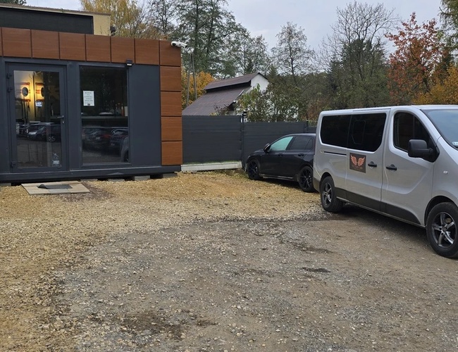 Victory Cars - Frente al edificio hay un autobús blanco. Al lado está estacionado un coche negro. En el fondo, árboles y una cerca gris.