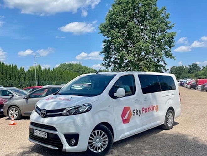 SkyParking - Un autobús blanco con logo está en un área de grava. Al fondo se ven árboles y coches aparcados. El tiempo es soleado.