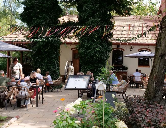 Parking Zielony Kompleks Dwór Ostoya – Ludzie siedzą w ogródku restauracyjnym. Otoczenie zdobią rośliny i parasole.