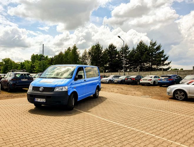 KTW Parking – Na wybrukowanym placu stoi niebieski bus. W tle parkują auta. Teren otaczają drzewa i ogrodzenie.