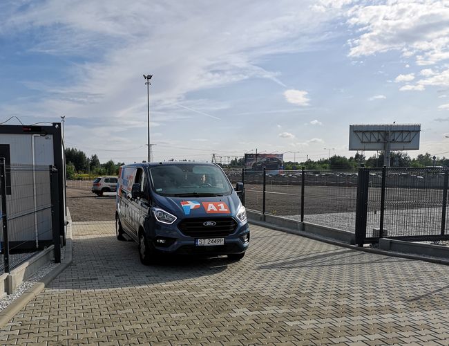 Parking A1 Rondo – Teren jest ogrodzony wysokim płotem z siatki. Na bruku stoi samochód dostawczy z logo A1.
