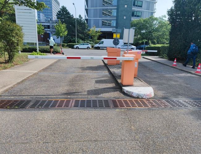 Parking FrankfurtValet24 - Szlaban wjazdowy blokuje drogę. W tle widać budynki i przyrodę. Po lewej stronie osoba jedzie na hulajnodze.